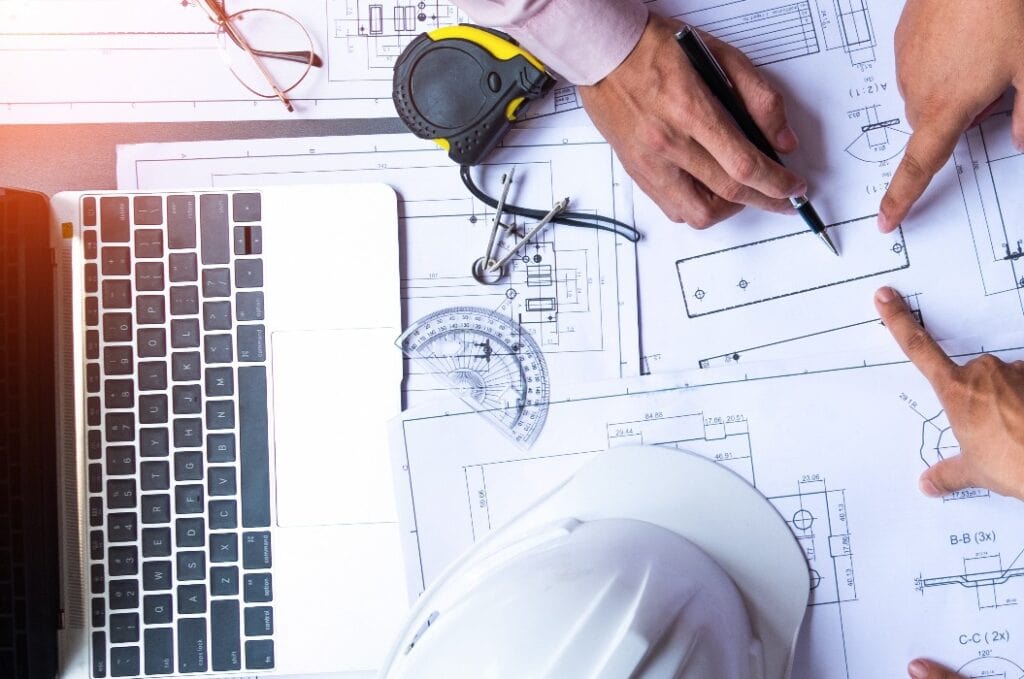 Die Hand eines Ingenieurs zeigt mit einem Stift auf einen Bauplan. Mit Laptop, Zirkel und Lineal zeichnet er den Entwurf für ein Bauprojekt.